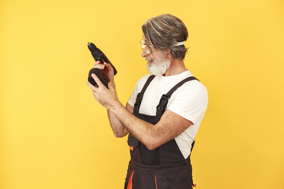 Mature handyman in overalls with safety glasses examining a power drill against a vibrant yellow backdrop.