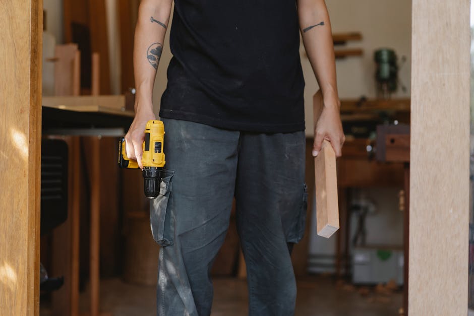 Unrecognizable craftsman in casual clothes standing with electric screwdriver and piece of wood while working in professional workshop