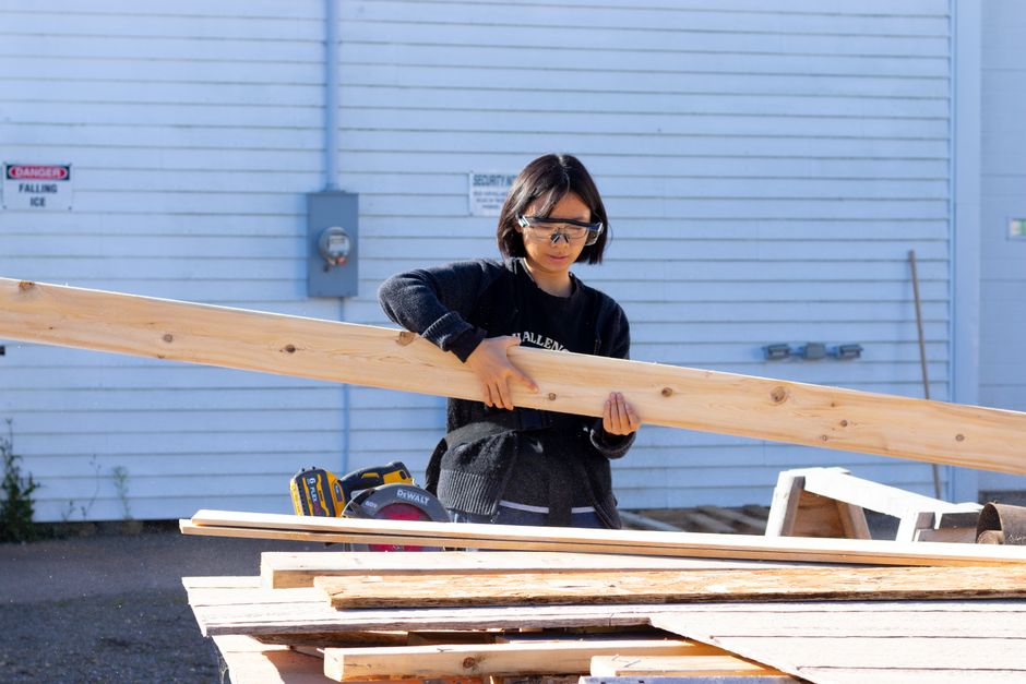A young woman using power tools and handling wood planks outdoors, wearing protective glasses.