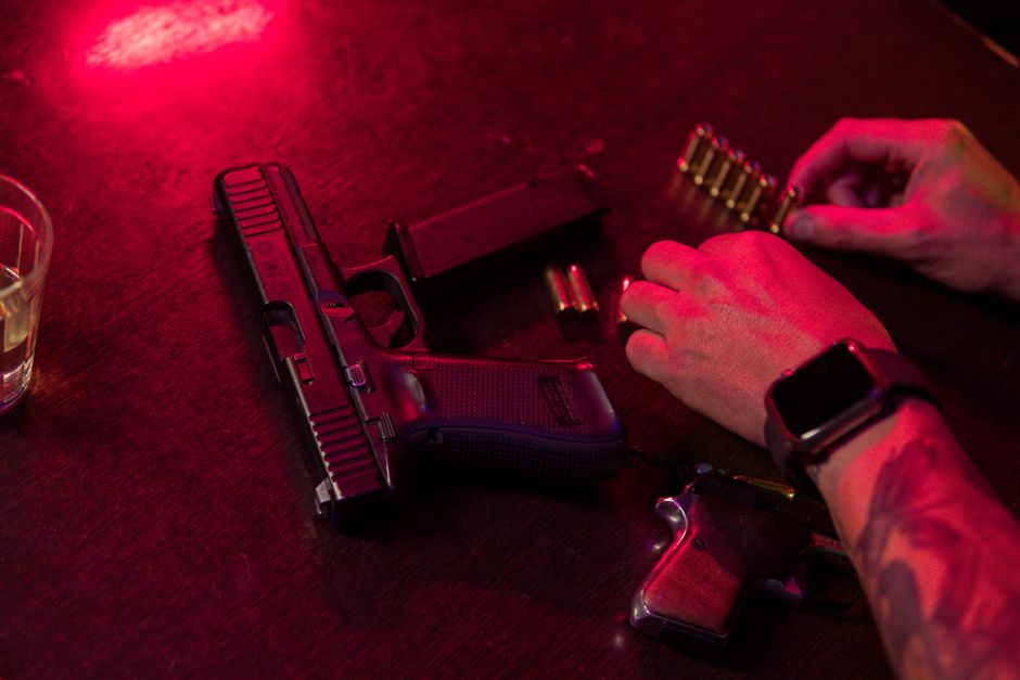 Dramatic close-up of a tattooed hand loading bullets beside a handgun and whiskey glass.