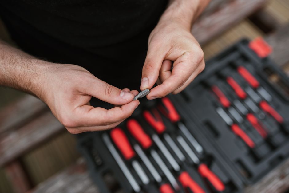 From above of crop anonymous master examining pin over tool box with screwdriver while working in professional workshop in soft daylight