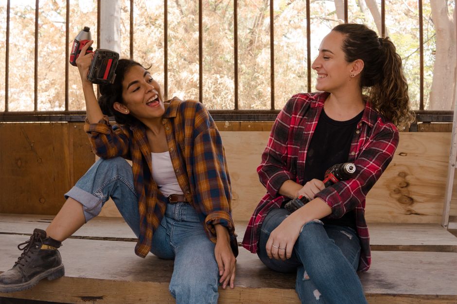 Two women in plaid shirts, laughing together in a workshop with power tools.