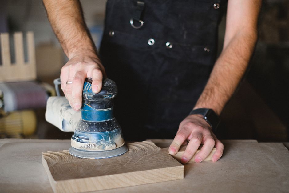 Unrecognizable male carpenter using random orbital sander while working with wooden detail at workbench in professional workshop on blurred background