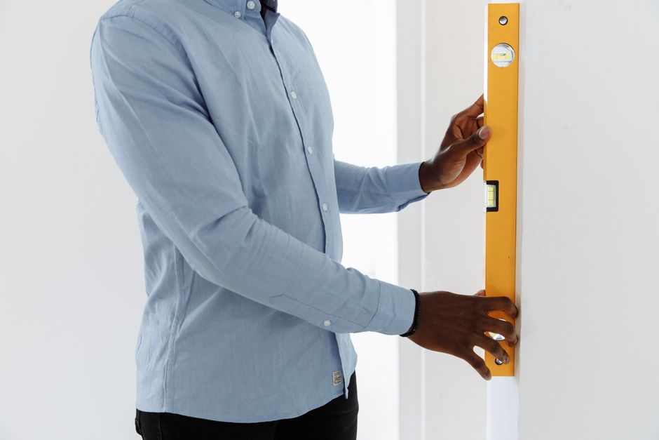 Adult man using a yellow spirit level against a white wall, wearing a blue shirt indoors.