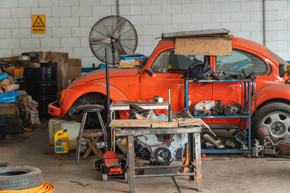 Classic red car being repaired in a cluttered garage setting, filled with tools and auto parts.