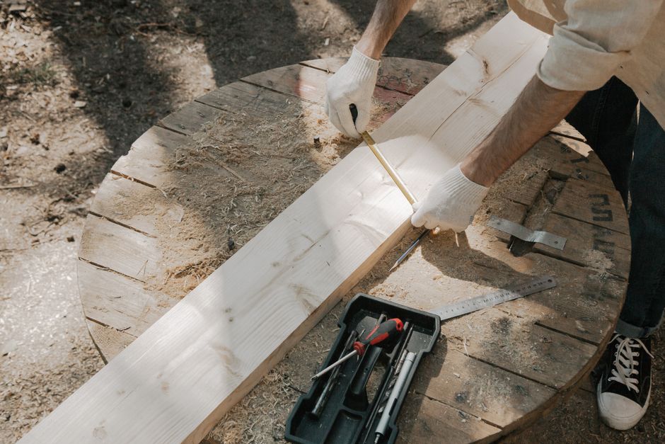 A carpenter in gloves measures a wooden plank on a round worktable with tools outdoors.