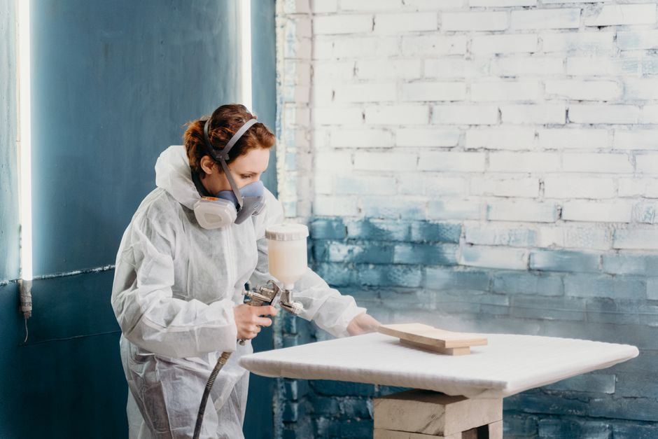 A female artisan wearing protective gear spray paints a wooden object indoors.