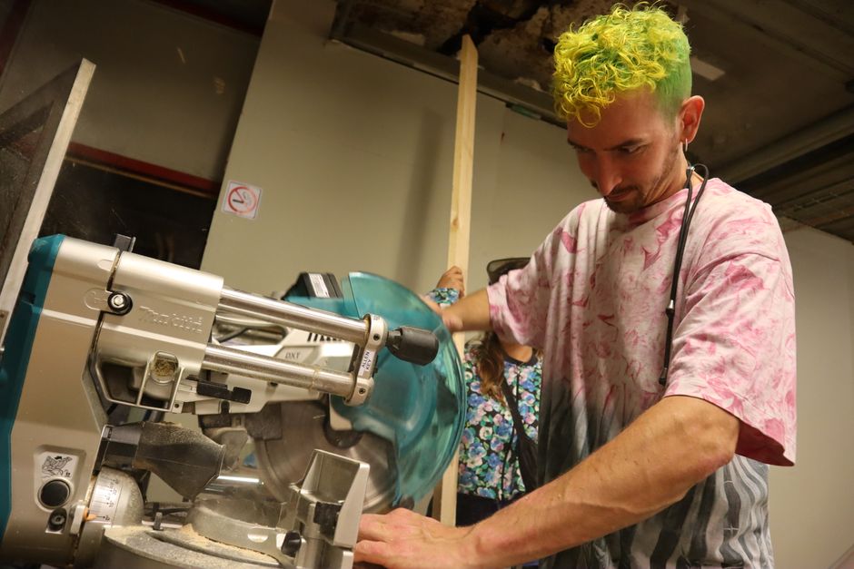 A focused young man with colorful hair using a miter saw in a workshop setting.