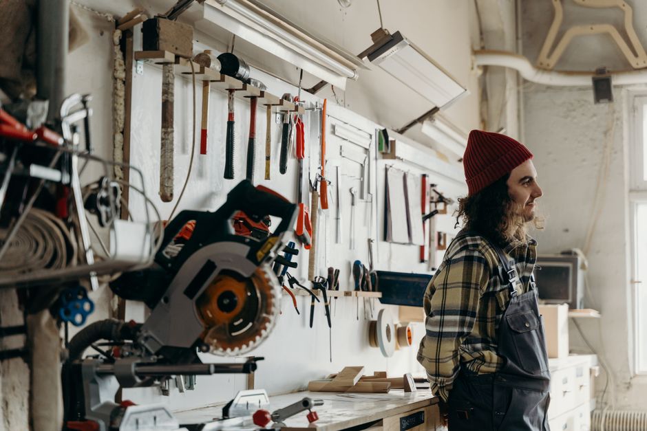 A man with long hair and beanie stands in a workshop with tools on the wall, reflecting a warm workspace atmosphere.