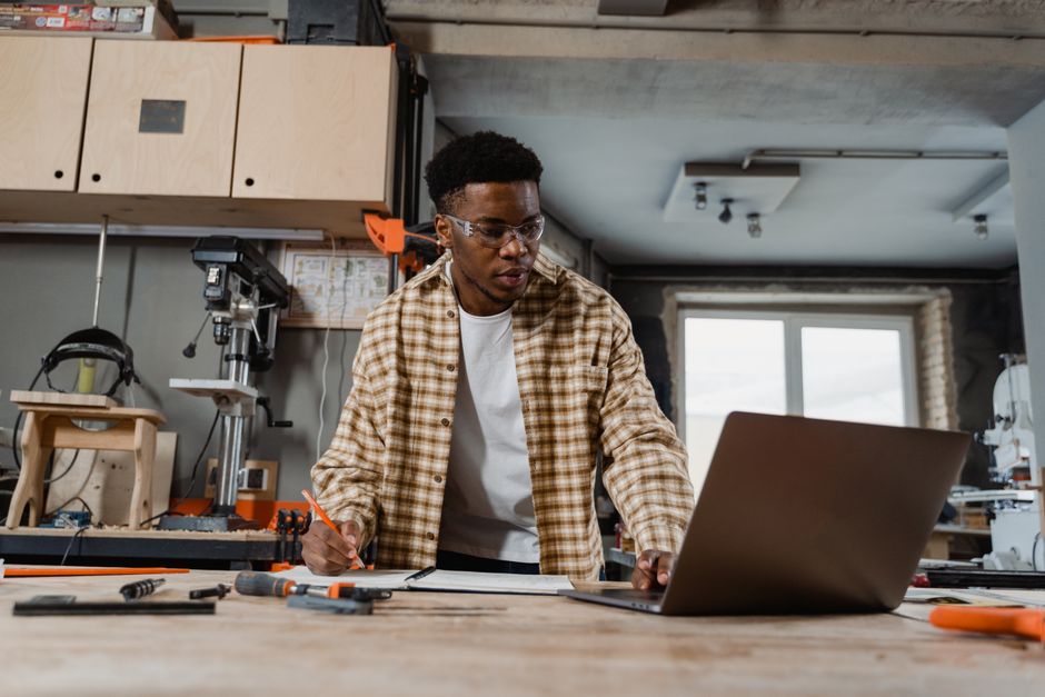 Carpenter using laptop and tools in a workshop, strategizing for a woodworking project.