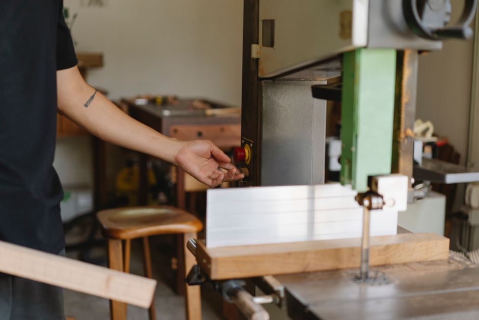 Crop anonymous tattooed carpenter pressing button on professional electric equipment while standing with wooden plank near table