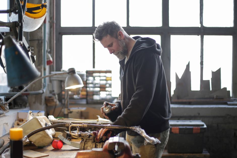 Side view of adult bearded mechanic in workwear using spray to prevent corrosion of metal detail while working at workbench in light workshop