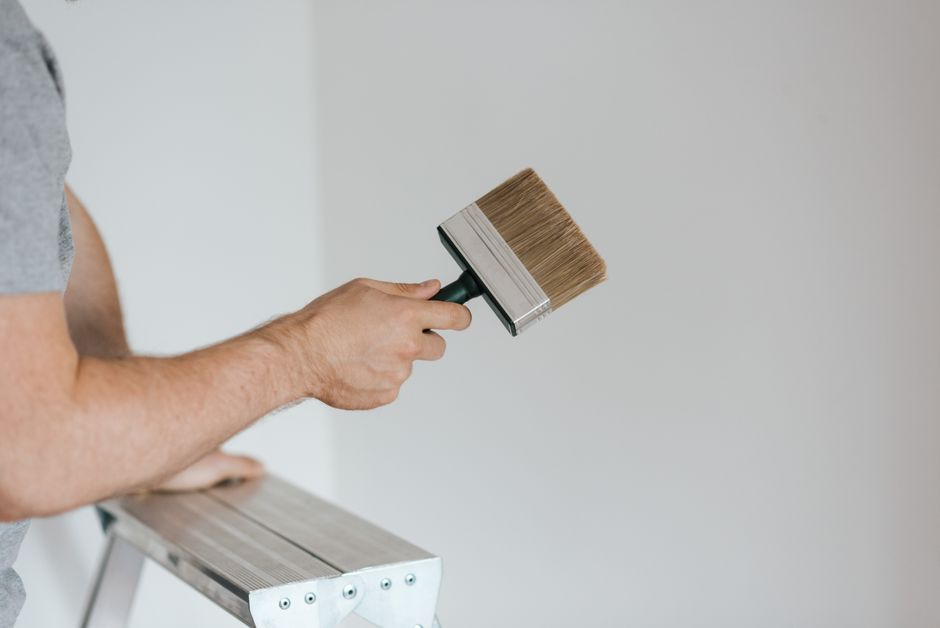 Side view of crop faceless painter in casual clothes holding paintbrush while leaning on stepladder on gray background in soft daylight