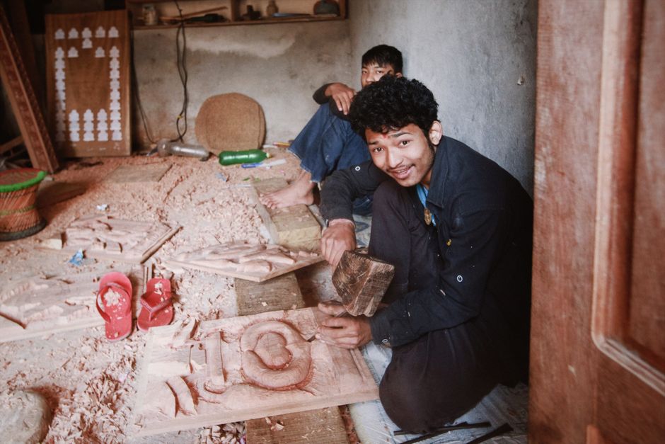 Two young artisans carving wood with traditional tools in an indoor workshop setting.