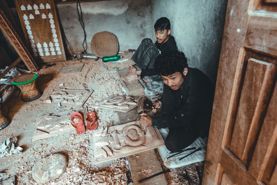 Two young artisans engaged in intricate wood carving in a small workshop, surrounded by tools and materials.