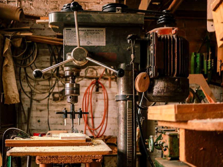 An old drill press in a workshop filled with tools and wooden planks.