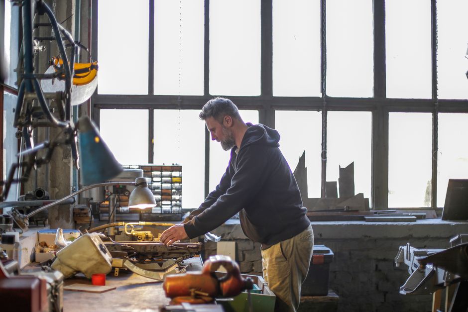 Side view of adult male bearded master in workwear standing near workbench and working with tools while fixing broken detail in workshop