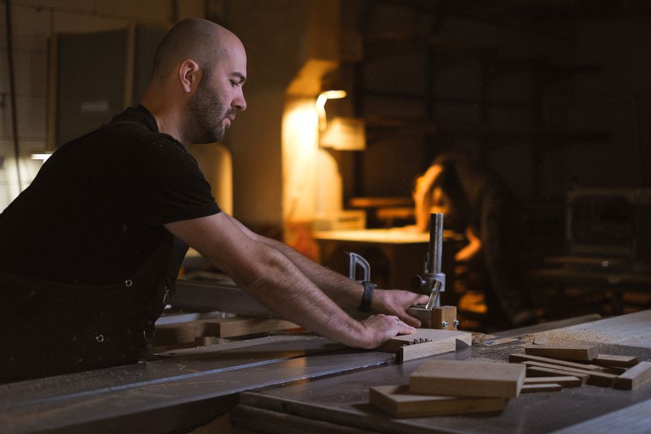Side view of male artisan cutting wooden plank with circular table saw while working in professional joinery with lamp on blurred background