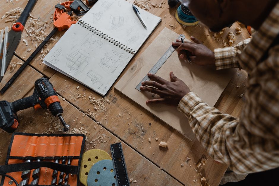 Woodworker measures timber amidst tools and sketches in a detailed workshop.
